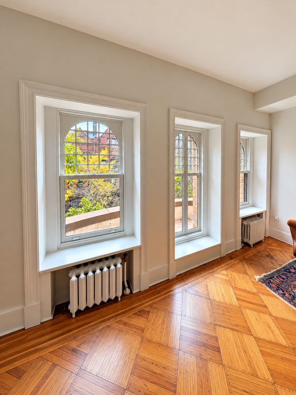 Interior wall with three tall white framed windows with arched heads and divided lites: a large double hung window, a narrower pair of casement sashes and a small double hung window, with a radiator under each double hung window, above a herringbone floor