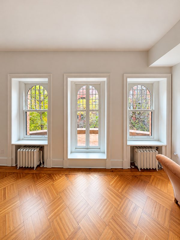 Interior wall with three tall white framed windows with arched heads and divided lites: a double hung window with a radiator beneath it on each side of a narrower pair of casement sashes that has no radiator, above a herringbone floor