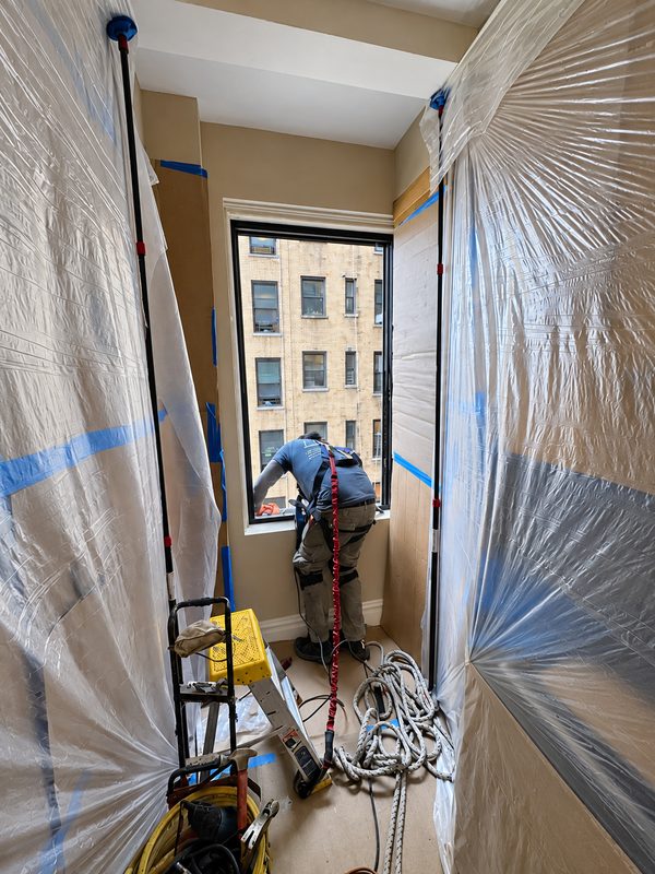 Installer at work at a window in an apartment, with plastic sheeting hung on both sides and protection taped to the floor