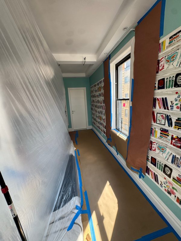 Apartment corridor with plastic sheeting along one wall, blue tape and floor protection beside a window and a painted wall