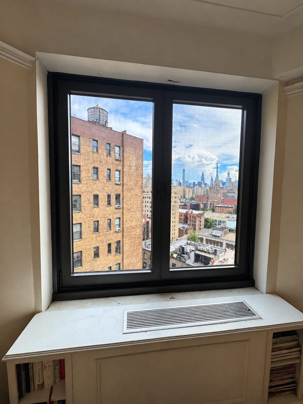 Wide new dark framed window with two sashes above a white window seat with a vent grille, looking at a brick building and the skyline