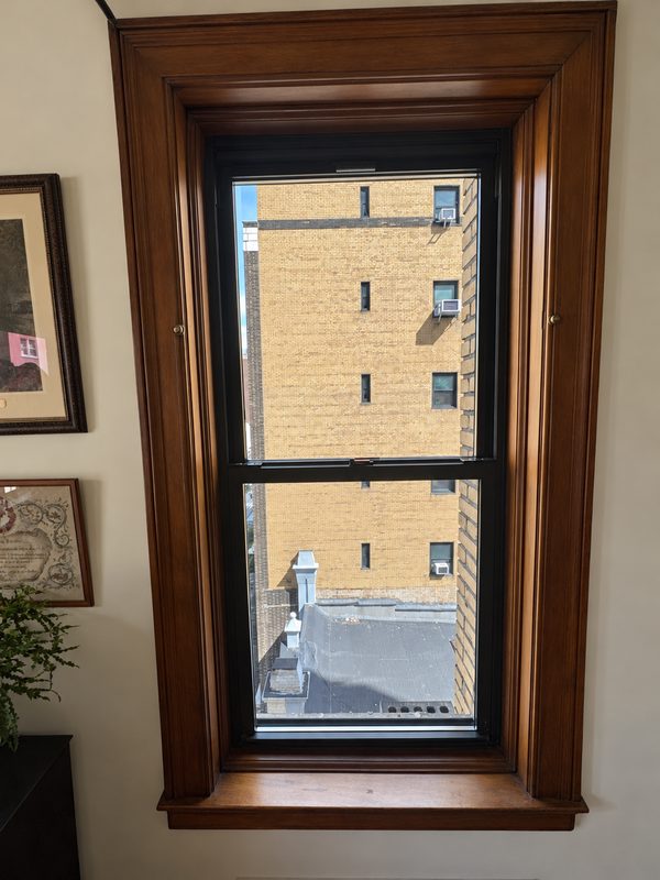 Tall new black framed double hung window in a dark wood casing, with framed pictures at the left
