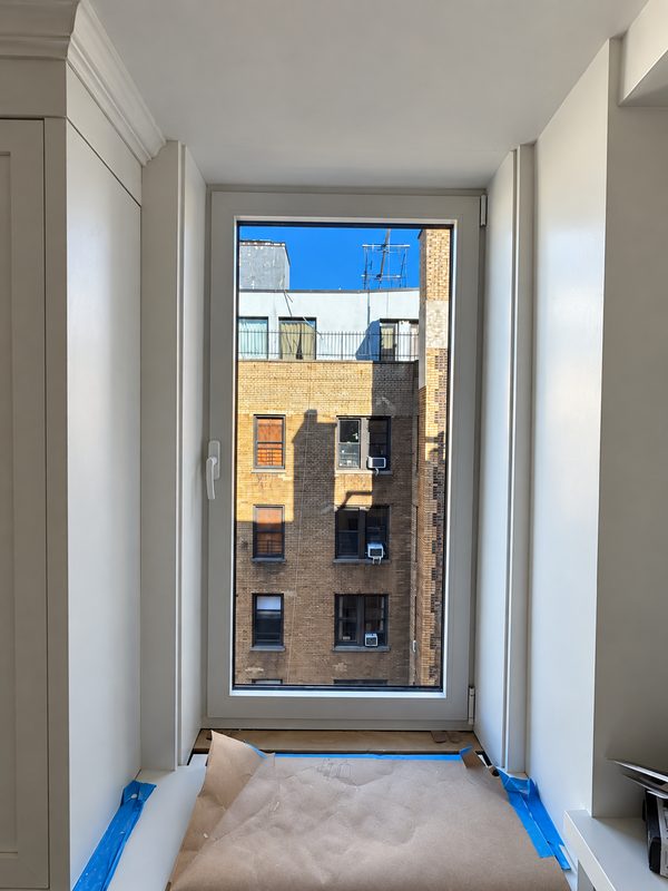 New white framed window in a deep white opening, with floor protection taped below, looking at a brick building