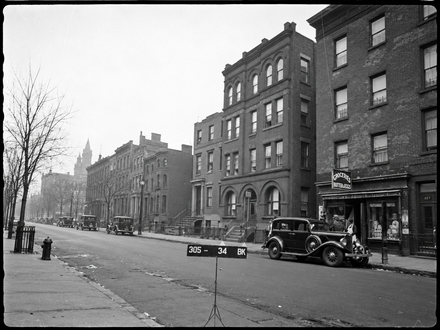 Archival black and white photograph of the street, with a four story building with arched windows beside a grocery store, an early automobile at the curb and a numbered placard in the roadway