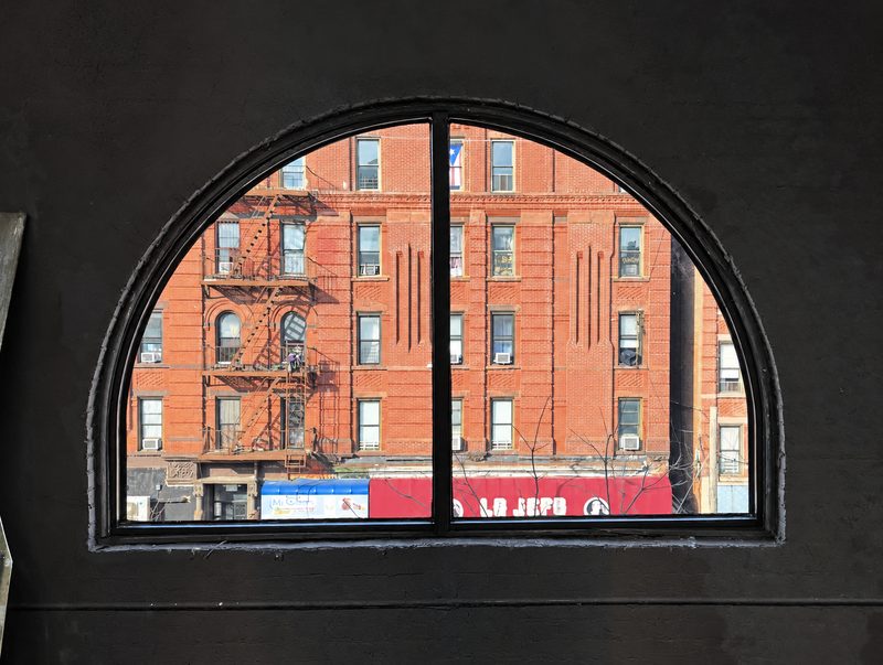 Arched window with a black frame and a center mullion seen from inside, with a red brick apartment building and fire escapes across the street in view