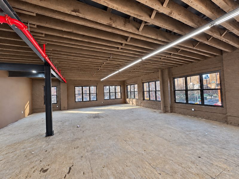 Open upper floor with exposed wood joists, a red pipe, a black steel column and new dark framed windows along the walls