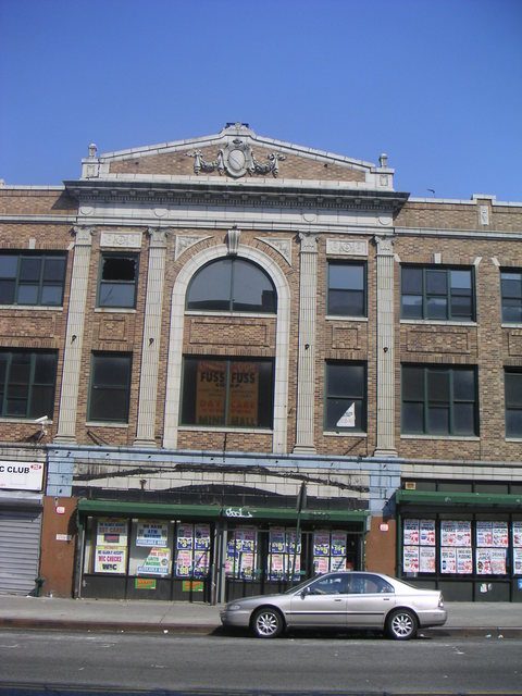 Older photograph of the central bay of the building, with its pediment and arched window above storefronts covered in posters, and a parked sedan
