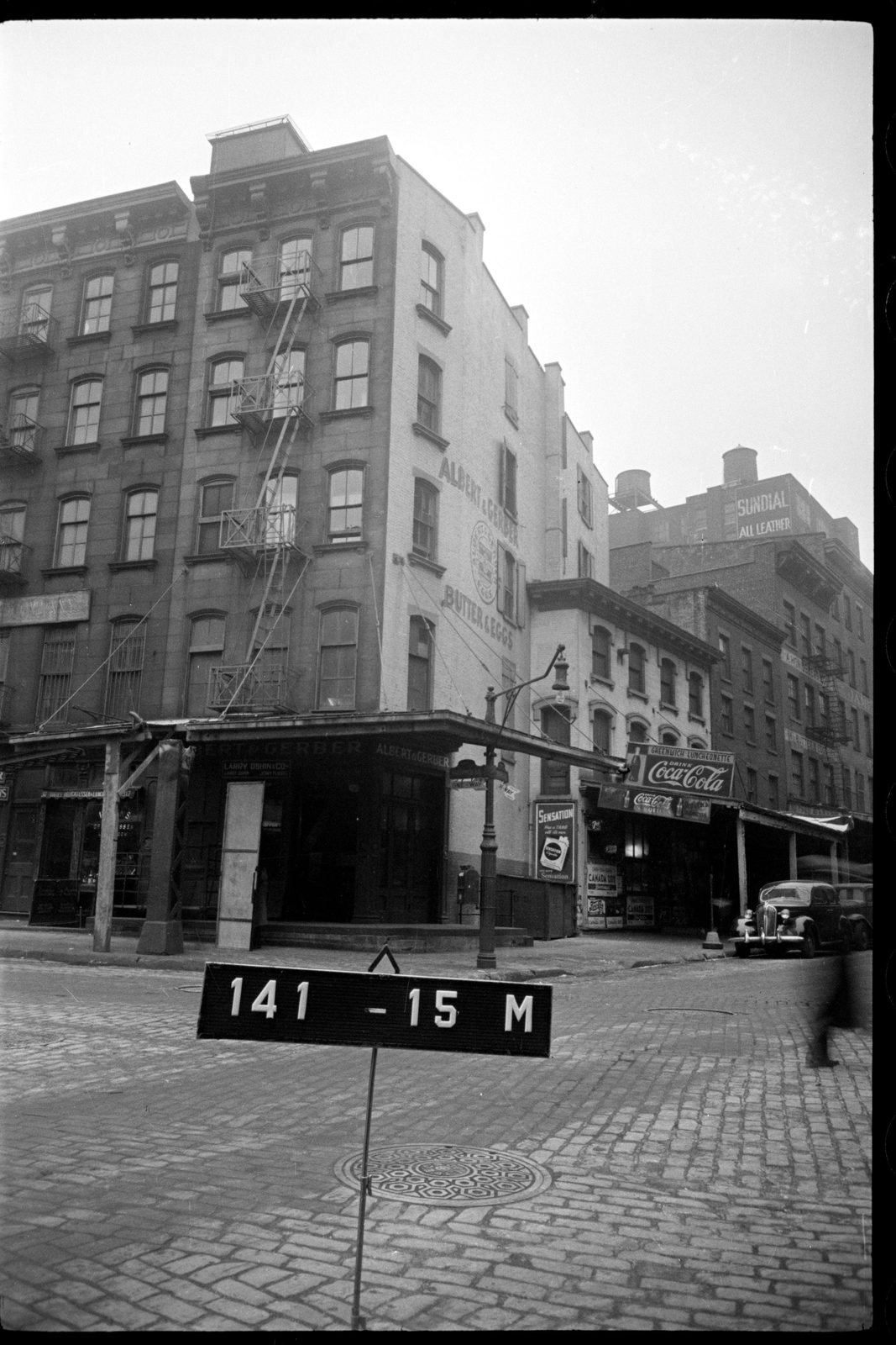 Archival black and white photograph of a street corner with cobblestones, a corner building with fire escapes and a numbered placard in the foreground