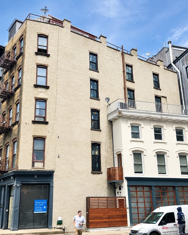 View up the tan side elevation from the street, with rows of new windows, a rooftop railing and a white van at the curb