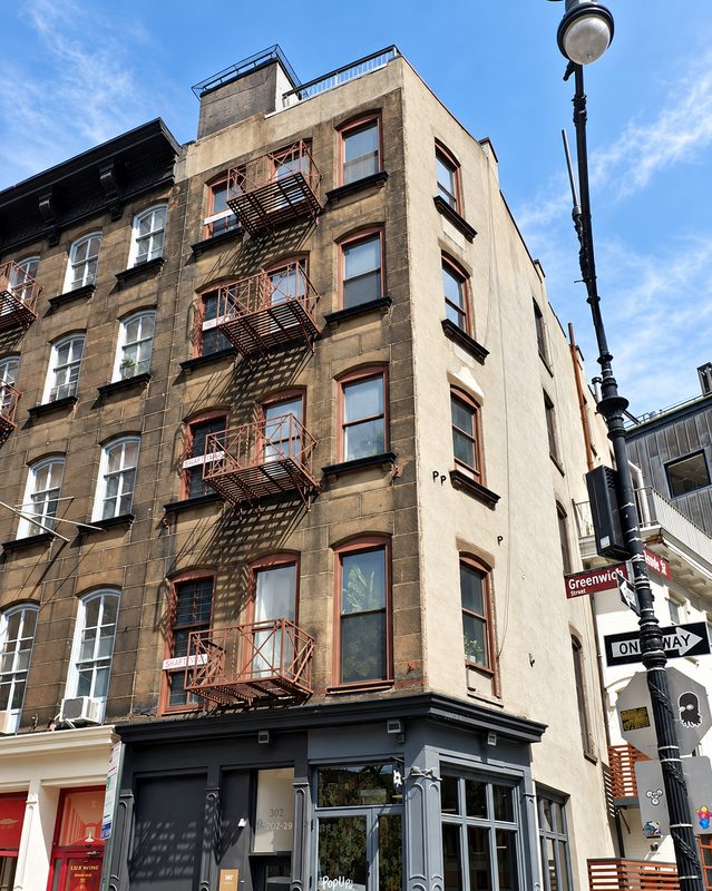 Upward view of the building at the street corner, with new brown framed windows, red fire escapes on the neighboring building, a street lamp and a one way sign
