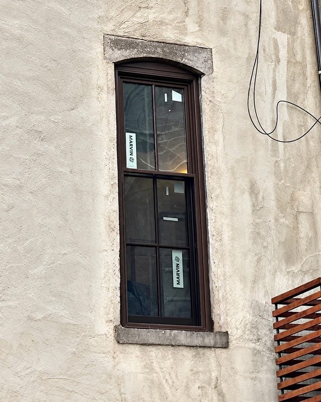 Close view of a tall new double hung window with a dark brown frame and an arched head, with Marvin labels on the glass, set in a stucco wall with stone trim