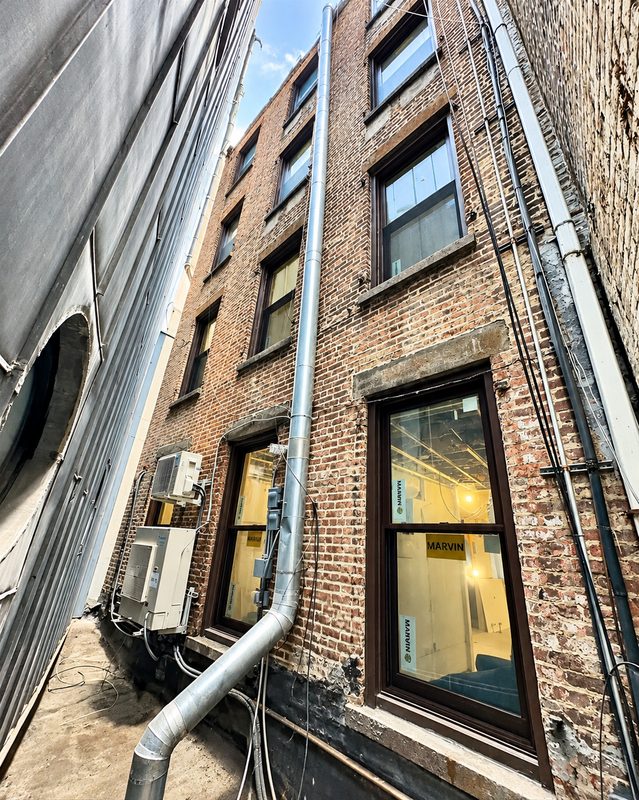 Rear elevation seen between buildings, with new brown framed windows in a brick wall beside pipes and mechanical units