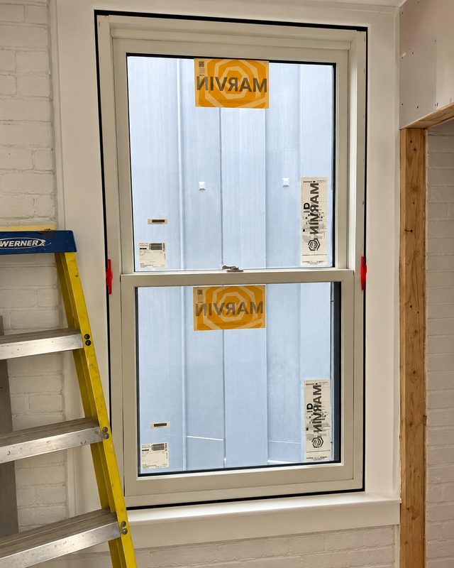 Inside view of a new double hung window with orange Marvin labels on the glass, beside a yellow ladder and a painted brick wall